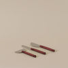 Three cheese knives with metallic silver blades and dark red handles, arranged diagonally on a plain beige background.