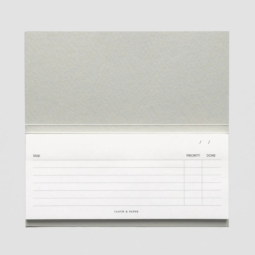 A single-page sticky note pad with a textured light gray cover and a white lined writing page showing columns for task, priority, and done, on a light gray background.
