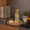 A clear glass carafe with a light wood lid sits on a round wooden tray on a dark countertop, alongside a small glass of lemon water, a metal lid, books, a potted plant, and an orange in a kitchen setting.
