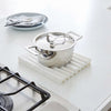 A stainless steel pot with a lid sits on a white ribbed silicone drying mat on a white kitchen countertop, with a gas stovetop burner partially visible in the foreground and a plate in the background.