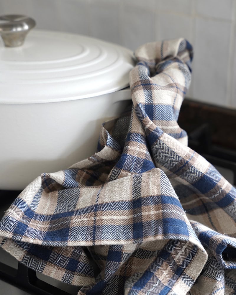 A plaid kitchen towel in blue, beige, and white draped over the side of a white enameled pot on a stovetop.