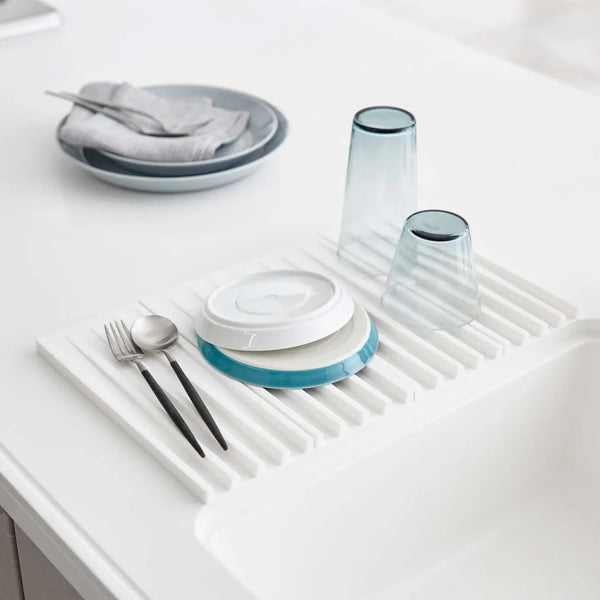 A white slotted sink drying mat shown on a white countertop, holding stacked plates, two translucent blue-gray tumblers, and utensils. 