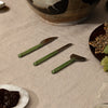Three green-handled cheese knives arranged on a beige linen tablecloth, with plates of food and a ceramic bowl partially visible in a styled dining setting.