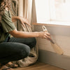 Person cleaning baseboards with a broom in a room with a window and curtain.