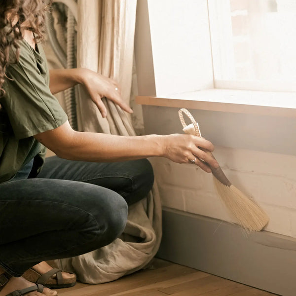 Person cleaning baseboards with a broom in a room with a window and curtain.