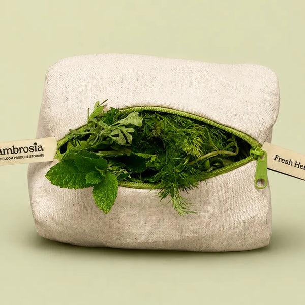 A square beige linen produce storage pouch with a green zipper opening, shown filled with fresh green herbs. Two paper tags are attached to the zipper, one reading 'Ambrosia' and the other partially visible as 'Fresh Herbs.