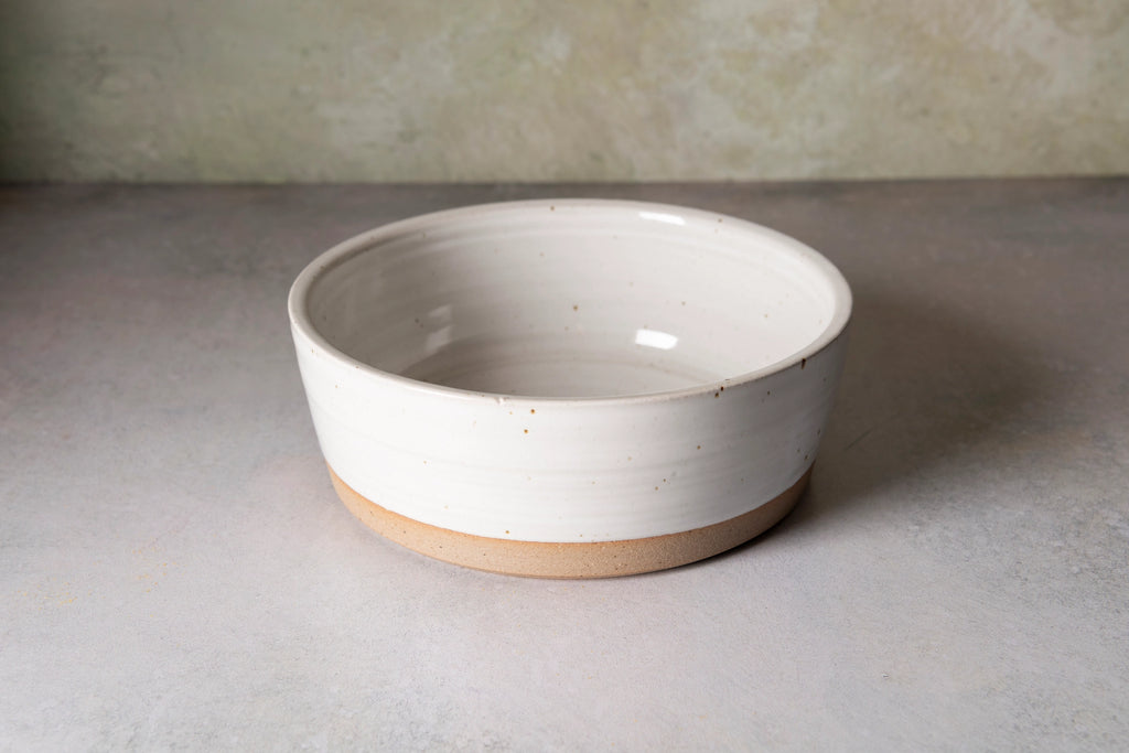 A handmade ceramic bowl with a glossy off-white glazed finish and a natural, speckled tan clay base, set against a neutral gray surface and textured beige wall.