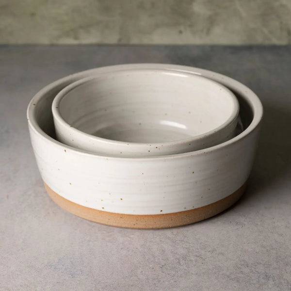 A set of two nested ceramic bowls with a speckled off-white glaze and a natural tan clay base, photographed on a textured gray surface.