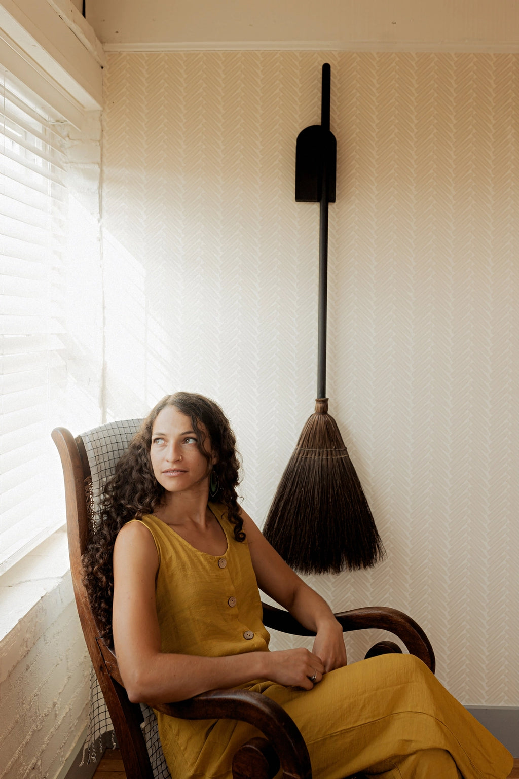 Woman in a yellow dress sitting in a chair next to a broom against a wall.