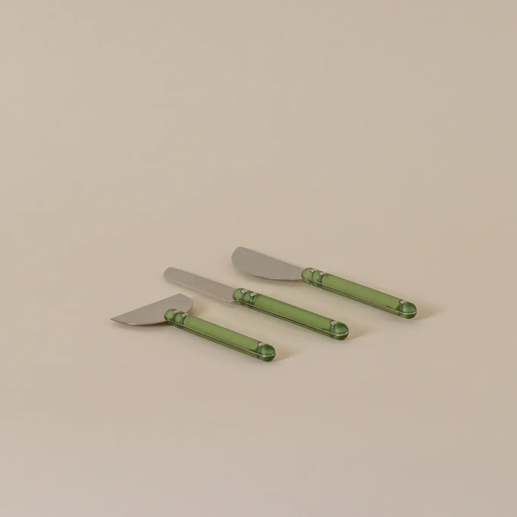 Three cheese knives with silver metal heads and translucent green handles, arranged diagonally on a plain beige background.