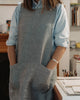 A person wearing a light blue button-up shirt and a gray linen apron with large front pockets, standing in a softly lit indoor room with shelves and desk items in the background.