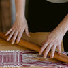 Close-up of a person's hands weaving a textile with burgundy and white threads over a wooden loom, with a wooden rolling pin resting across the woven area.