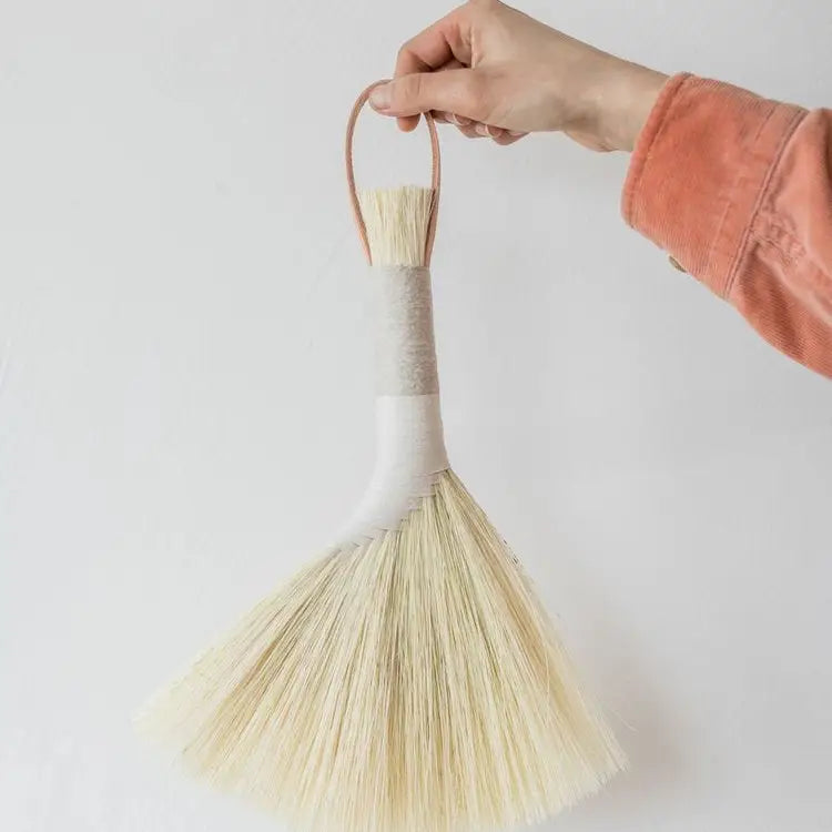 Hand holding a broom with a white handle and beige bristles against a plain background