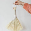 Hand holding a broom with a white handle and beige bristles against a plain background