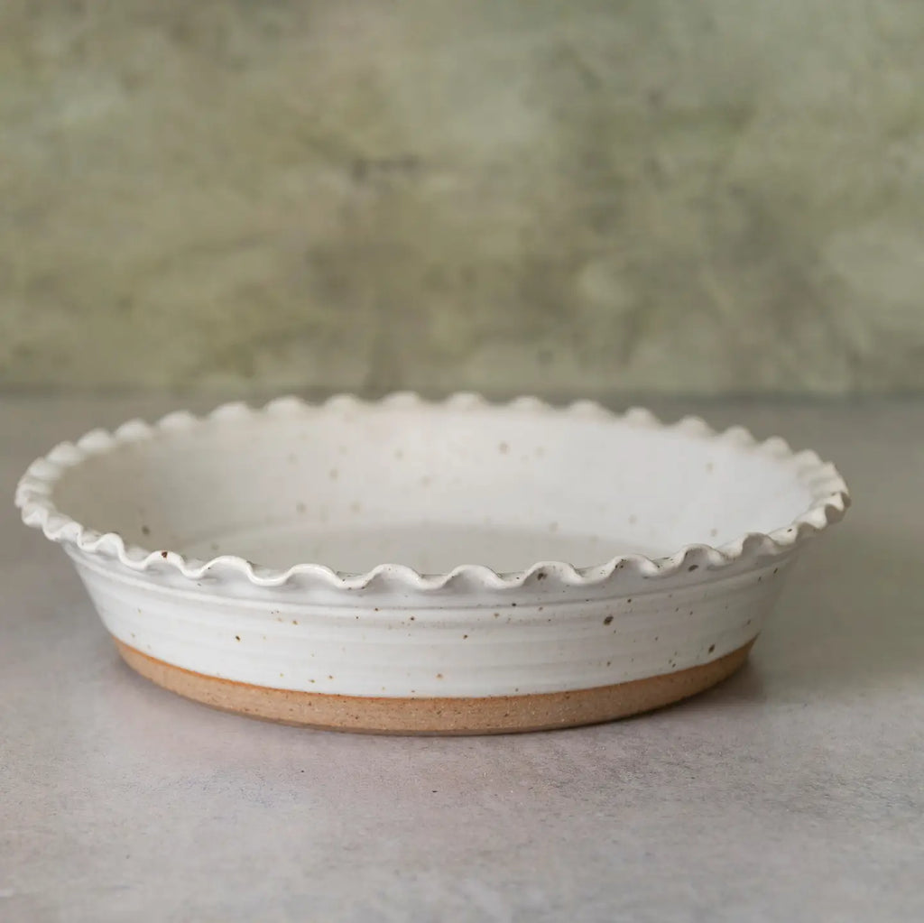 A round ceramic pie dish with a scalloped fluted rim, featuring a speckled off-white glazed exterior and a natural light brown clay base, set against a neutral textured background.