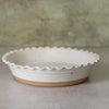 A round ceramic pie dish with a scalloped fluted rim, featuring a speckled off-white glazed exterior and a natural light brown clay base, set against a neutral textured background.