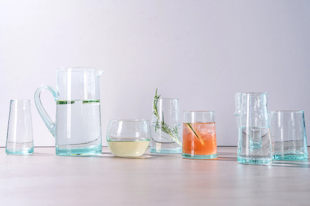 A set of clear glass drinking vessels with a light aqua blue tinted base, including a pitcher, tumbler, glasses, and a tumbler with a glass, arranged on a light wooden surface against a pale gray background.