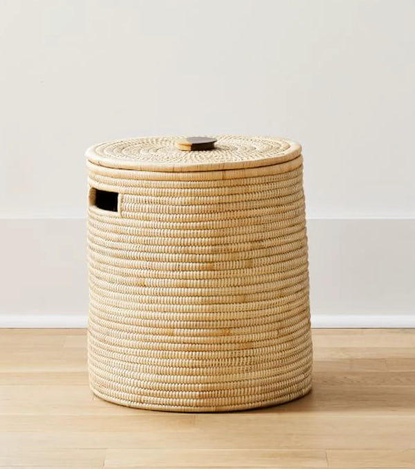 A natural woven rattan or seagrass storage basket with a matching lid and a small side cutout, standing on a light wood floor against a white wall.