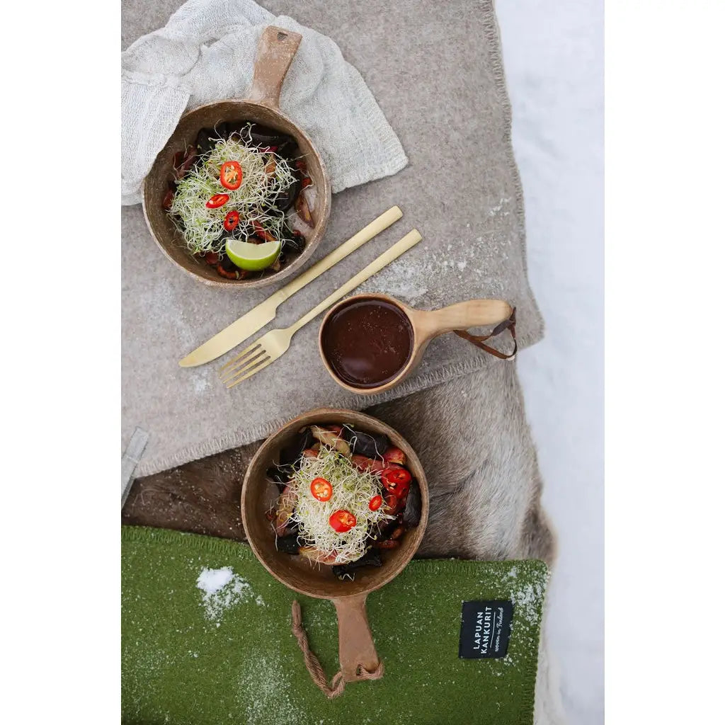 Two brown wooden bowls filled with a salad, arranged on a beige blanket with a knife, fork, and a small wooden cup of sauce. 