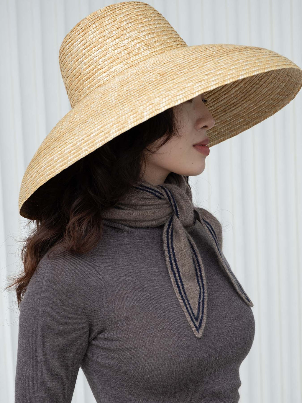 Person wearing a wide-brimmed straw hat and gray top with a scarf against a white background
