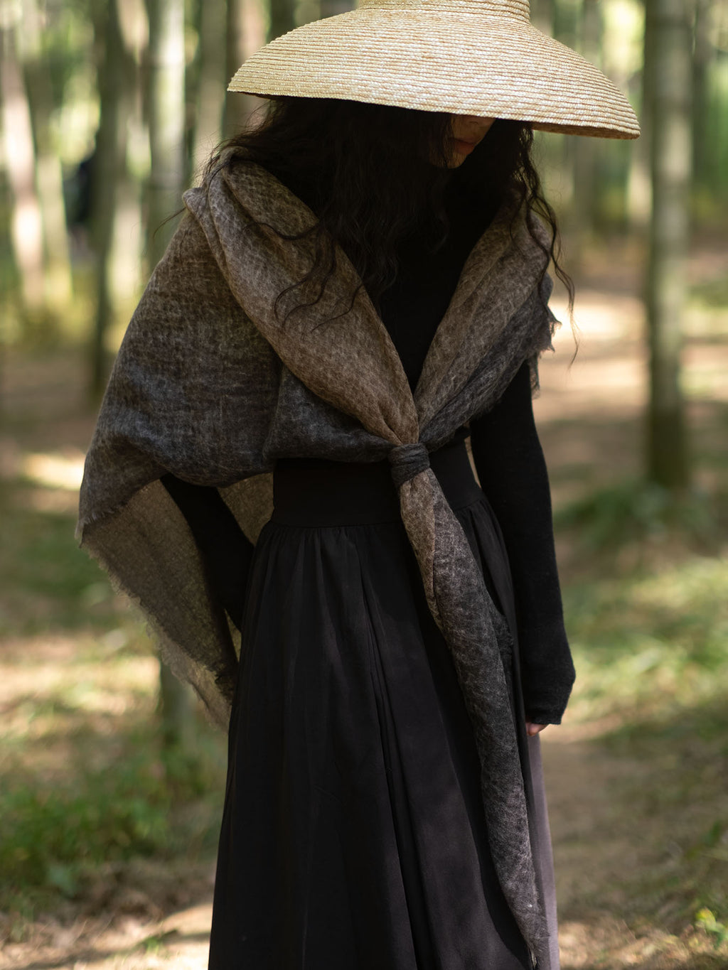 A person standing in a wooded outdoor setting wearing a wide-brimmed straw hat, a long black dress, and a large shawl draped over the shoulders in earthy brown and gray tones.