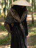 A person standing in a wooded outdoor setting wearing a wide-brimmed straw hat, a long black dress, and a large shawl draped over the shoulders in earthy brown and gray tones.