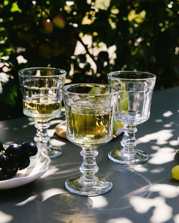 Three clear glass goblets with a drink on a reflective surface with greenery in the background