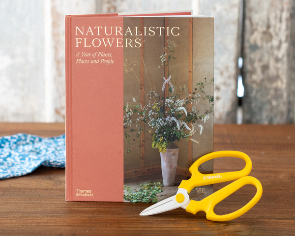 The book 'Naturalistic Flowers: A Year of Plants, Places and People' by Thames & Hudson, displayed on a wooden table next to a pair of yellow-handled scissors.