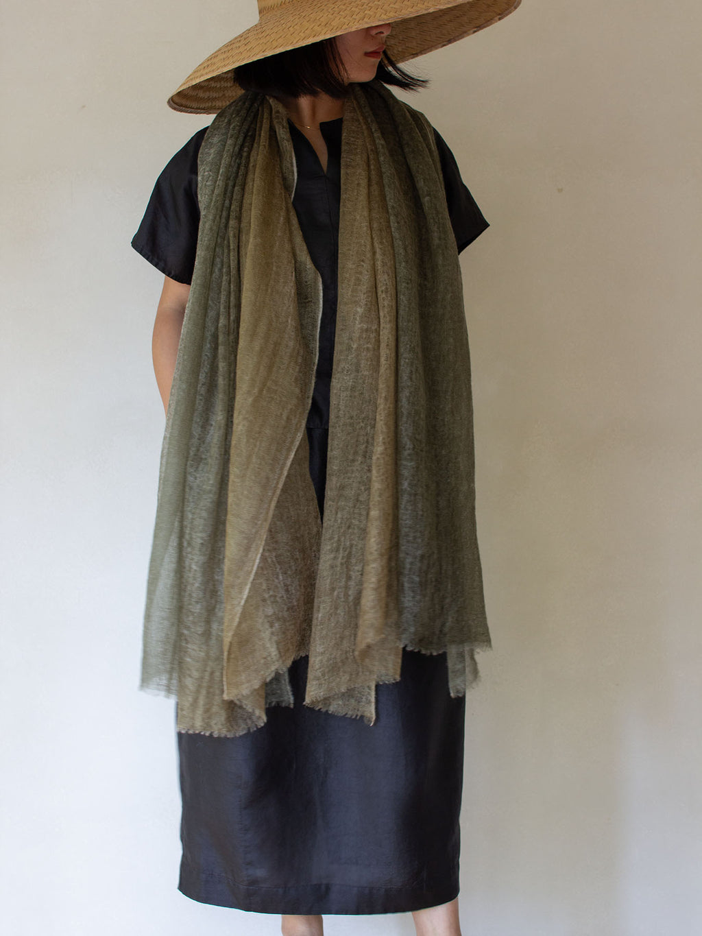 A person wearing a wide-brim straw hat, a black short-sleeve top, and a black skirt, with a long sheer olive-brown scarf draped over the shoulders against a plain light wall.