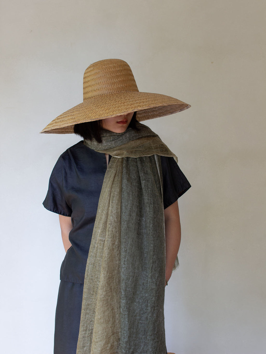A person wearing a large straw sun hat, a dark navy short-sleeve top, and a textured olive green scarf, standing against a plain off-white wall.
