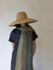 A person wearing a large straw sun hat, a dark navy short-sleeve top, and a textured olive green scarf, standing against a plain off-white wall.