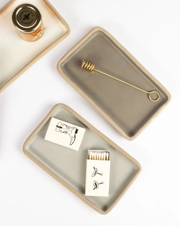 Two ceramic trays with decorative items including a bottle, matches, and a small tool on a white background.