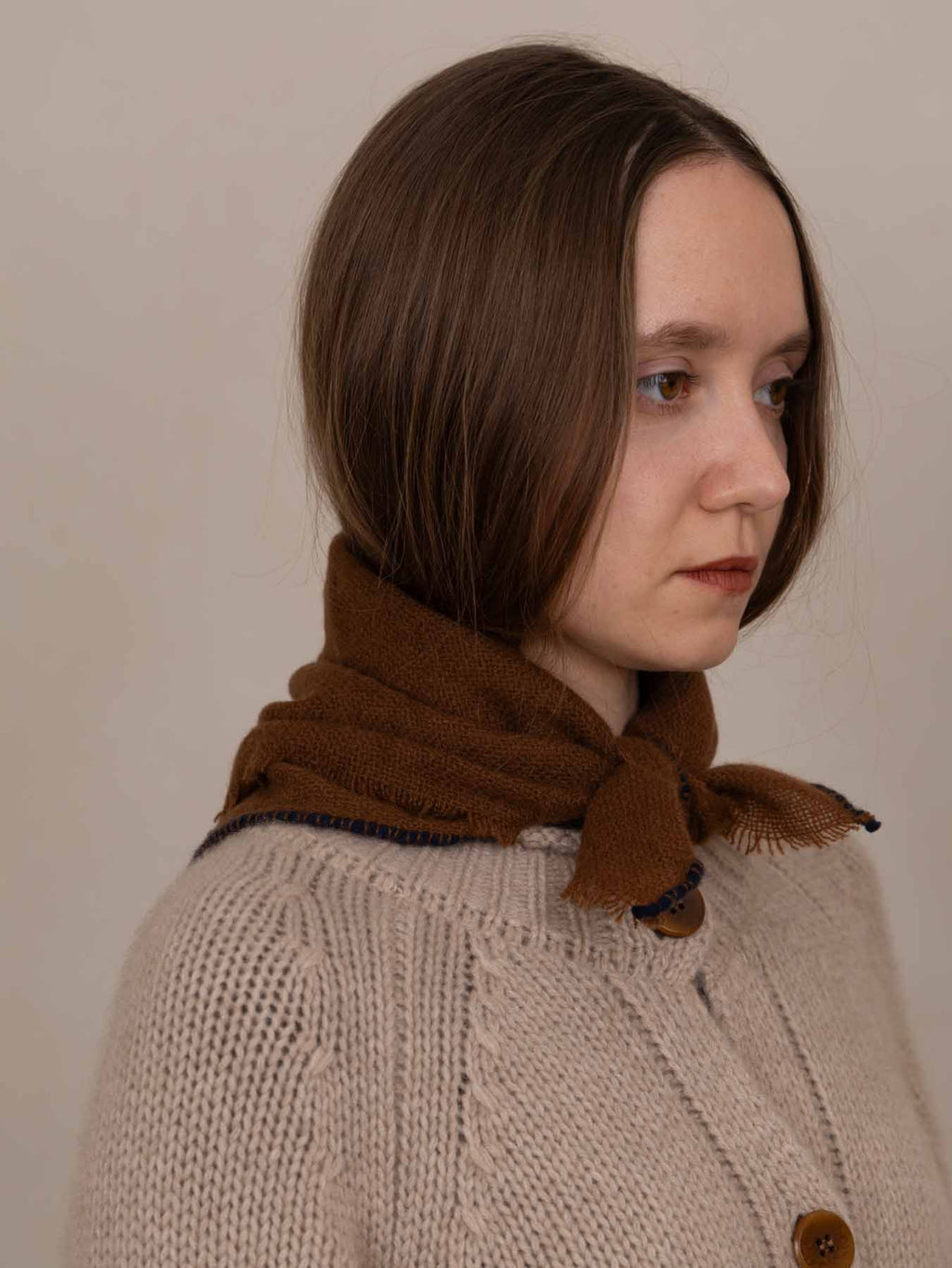 A woman with shoulder-length brown hair is shown in profile wearing a chunky knit beige cardigan and a brown scarf with dark blue trim, set against a plain beige background.