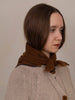 A woman with shoulder-length brown hair is shown in profile wearing a chunky knit beige cardigan and a brown scarf with dark blue trim, set against a plain beige background.