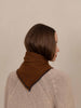 A woman shown from the back wearing a brown knit cowl neck scard over a cream cable-knit sweater, set against a plain beige background.