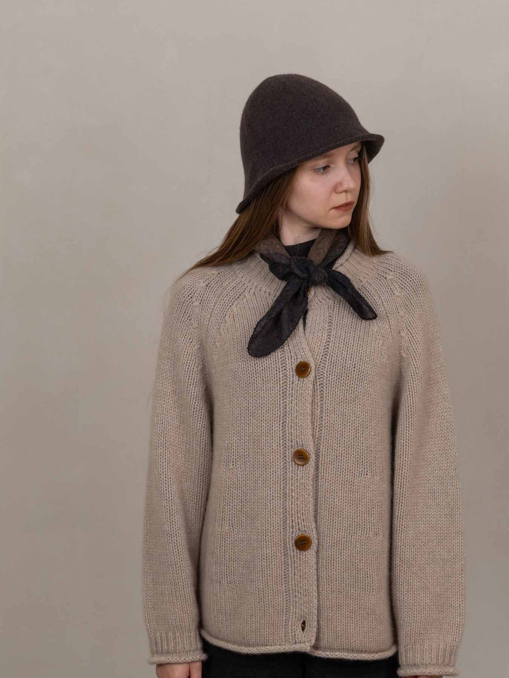 Person wearing a beige knitted cardigan with brown buttons, a dark hat, and a brown scarf against a plain background.