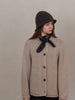 Person wearing a beige knitted cardigan with brown buttons, a dark hat, and a brown scarf against a plain background.
