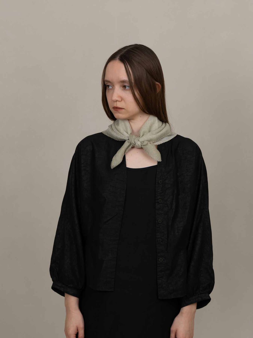 A model wearing a black textured cardigan with voluminous balloon sleeves and a light sage green knotted scarf, posed against a neutral beige studio background.