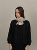 A model wearing a black textured cardigan with voluminous balloon sleeves and a light sage green knotted scarf, posed against a neutral beige studio background.