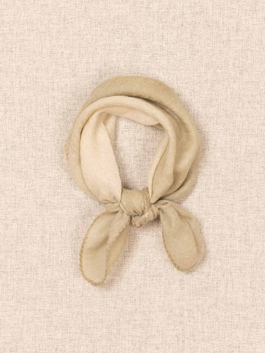 A beige knotted scarf made of soft cashmere fabric, displayed on a textured beige linen-like background.