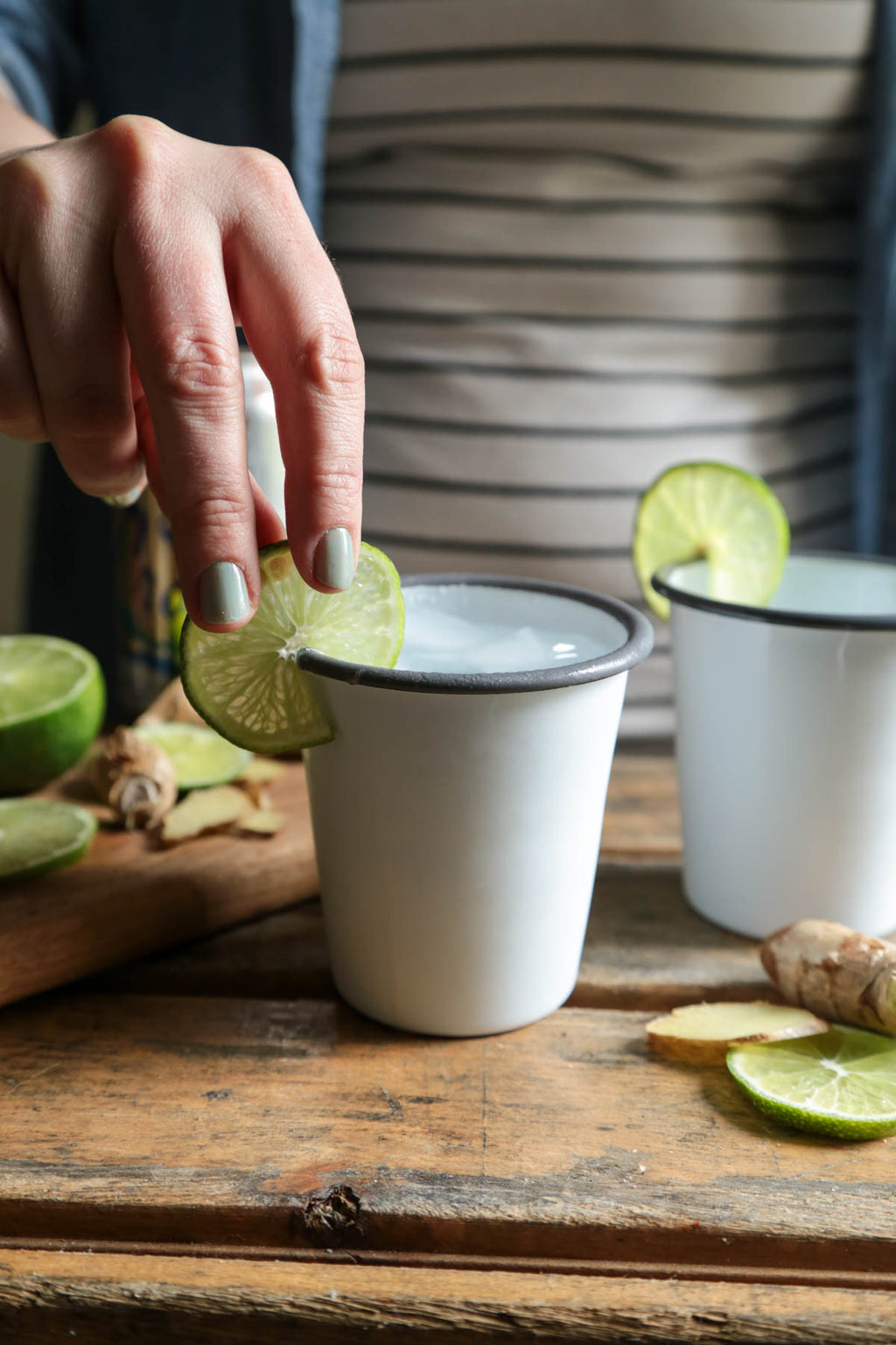 Person adding lime to a white cup on a wooden surface with limes and ginger around.