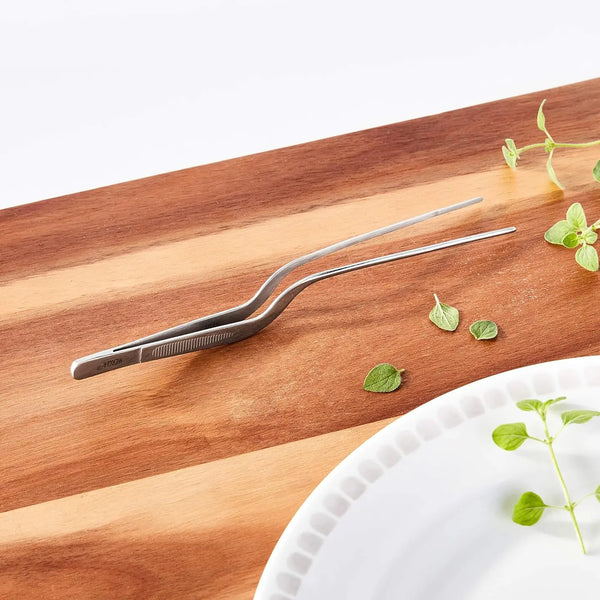 Metallic tongs on a wooden cutting board with green leaves.