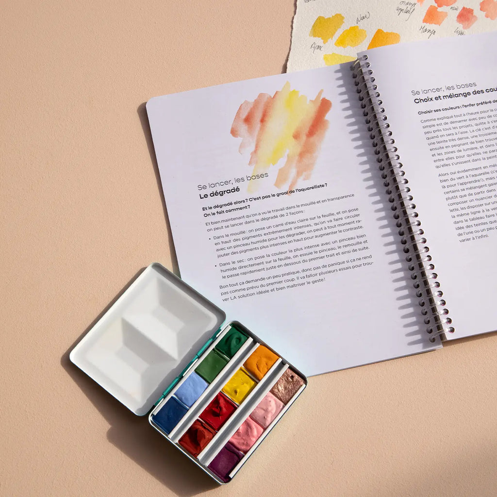 Open book with watercolor illustrations and a small watercolor set on a beige surface