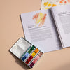 Open book with watercolor illustrations and a small watercolor set on a beige surface