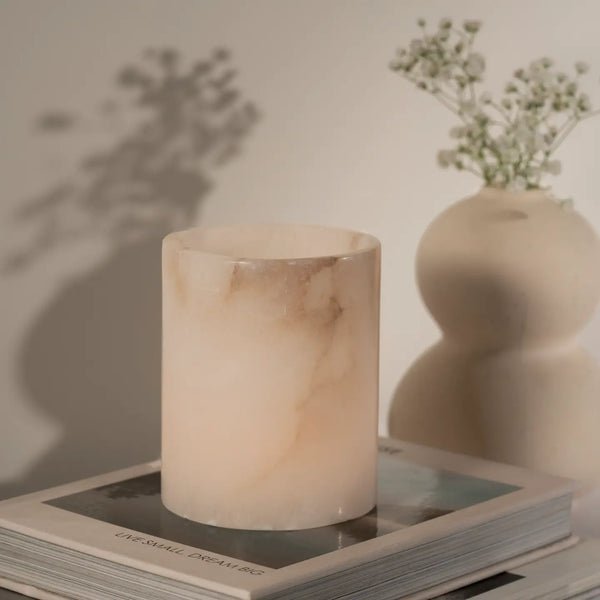 A pale blush pink marble candle holder or candle vessel with soft beige veining, styled on top of books with a blurred cream vase of small flowers in the background.