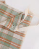 A close-up of a kitchen cloth with a muted plaid pattern in sage green, terracotta orange, and cream, featuring a cream canvas strap.