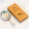 A stainless steel julep strainer with a perforated bowl and handle, shown next to its brown kraft paper packaging box on a light marble surface.