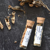 Two glass vials with cork lids containing dried seeds on a dark surface.