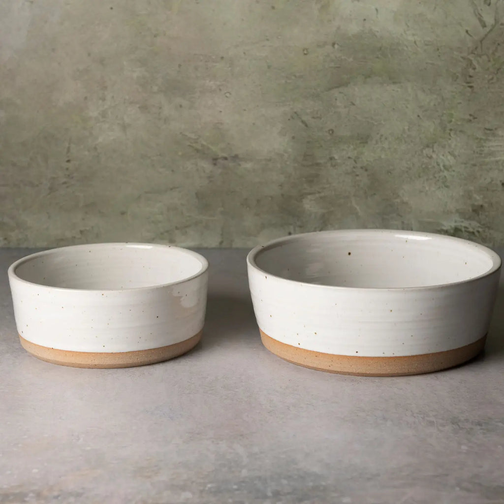 Two round ceramic bowls with a speckled off-white glaze and a natural tan unglazed base, displayed on a gray surface against a textured gray-green wall.