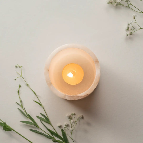 A lit tealight candle in a round, light beige stone holder, styled with delicate green and white baby's breath flowers on a neutral light gray background.